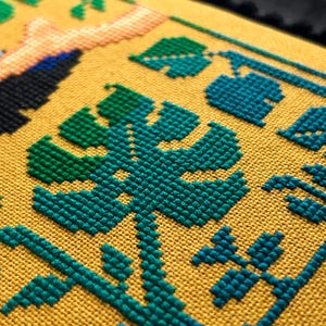 以下が含まれることがあります： 黄色の生地に緑と青のクロスステッチ刺繍が施されたクローズアップ。デザインは、さまざまな葉や植物を特徴としています。