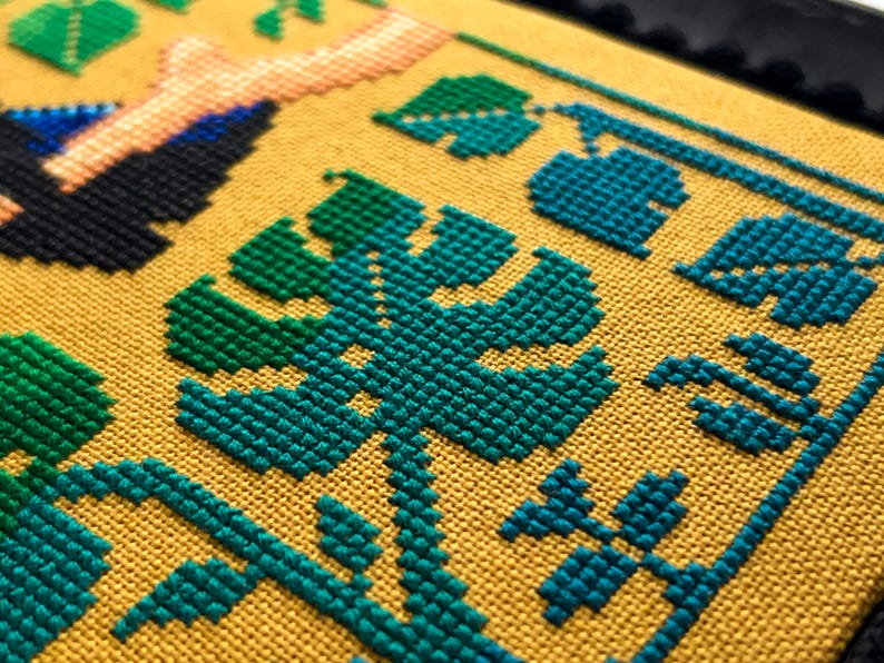 以下が含まれることがあります： 黄色の生地に緑と青のクロスステッチ刺繍が施されたクローズアップ。デザインは、さまざまな葉や植物を特徴としています。