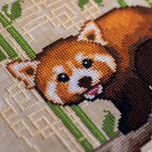 Pu&ograve; includere: Punto croce di un panda rosso con pelliccia arancione, marrone e bianca e occhi neri. Il panda &egrave; circondato da steli di bamb&ugrave; verdi e uno sfondo beige con un motivo geometrico. Il punto croce &egrave; dettagliato.