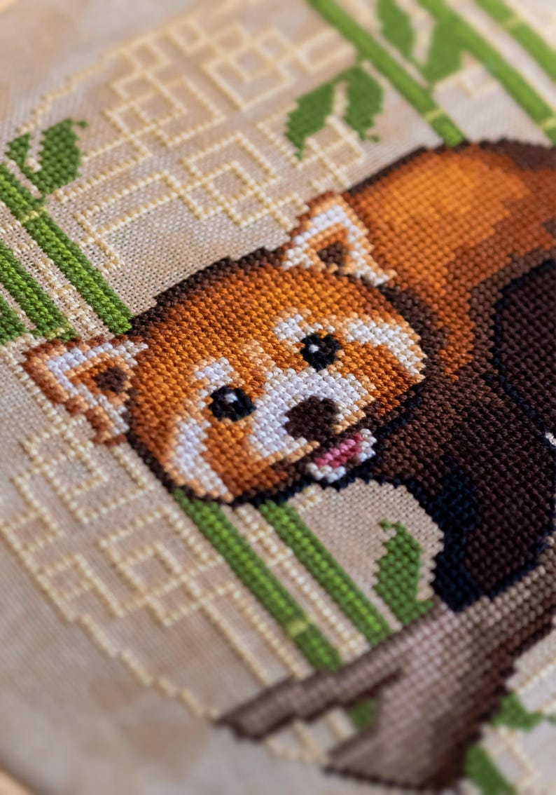 Pu&ograve; includere: Punto croce di un panda rosso con pelliccia arancione, marrone e bianca e occhi neri. Il panda &egrave; circondato da steli di bamb&ugrave; verdi e uno sfondo beige con un motivo geometrico. Il punto croce &egrave; dettagliato.