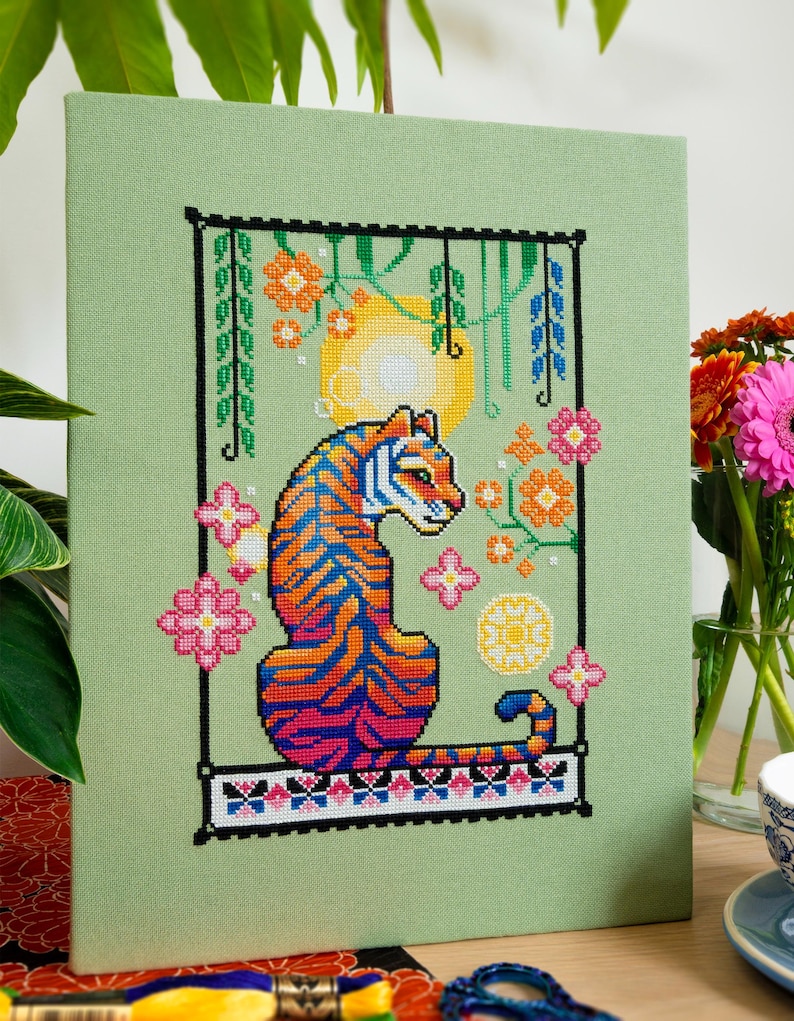 May include: Cross-stitch art of a tiger with orange and blue stripes, set against a light green background. The design includes flowers, a sun, and a decorative border. The artwork is framed and displayed on a wooden surface.