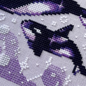 May include: Close-up of a cross-stitch pattern featuring two orcas, one in black and white and the other in shades of purple and white, swimming against a white background.
