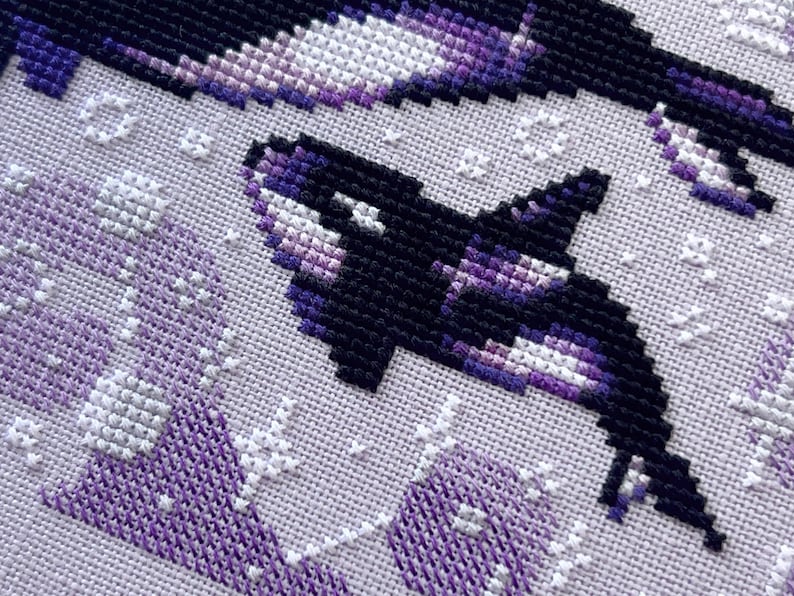 May include: Close-up of a cross-stitch pattern featuring two orcas, one in black and white and the other in shades of purple and white, swimming against a white background.