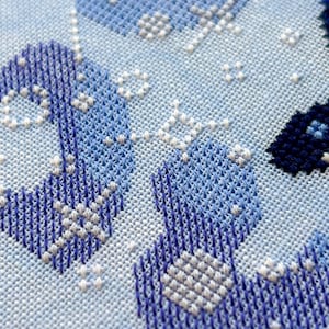 May include: Close-up of a cross-stitch design in shades of blue and white. The needlework features geometric patterns and shapes, creating a textured appearance on a light blue fabric. The stitches are detailed and precise.