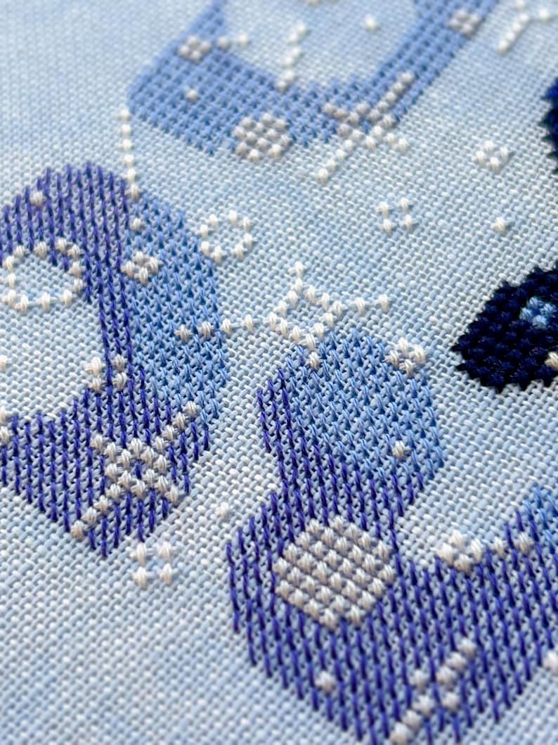 May include: Close-up of a cross-stitch design in shades of blue and white. The needlework features geometric patterns and shapes, creating a textured appearance on a light blue fabric. The stitches are detailed and precise.