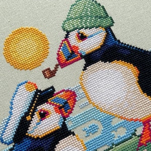 May include: Two puffins, one wearing a green hat and the other a blue and white sailor hat, are embroidered on a light green background. The puffin wearing the green hat is holding a pipe in its beak. The background also features a yellow sun and blue and white waves.