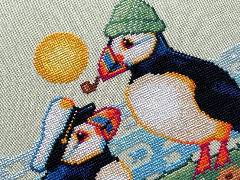 May include: Two puffins, one wearing a green hat and the other a blue and white sailor hat, are embroidered on a light green background. The puffin wearing the green hat is holding a pipe in its beak. The background also features a yellow sun and blue and white waves.