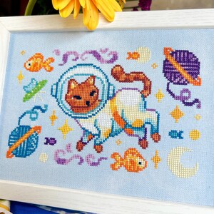 May include: Cross-stitch art featuring a cat in an astronaut suit, surrounded by space-themed elements like planets, fish, and stars. The artwork is set against a light blue background and framed in a white wooden frame. The cat is brown and white.