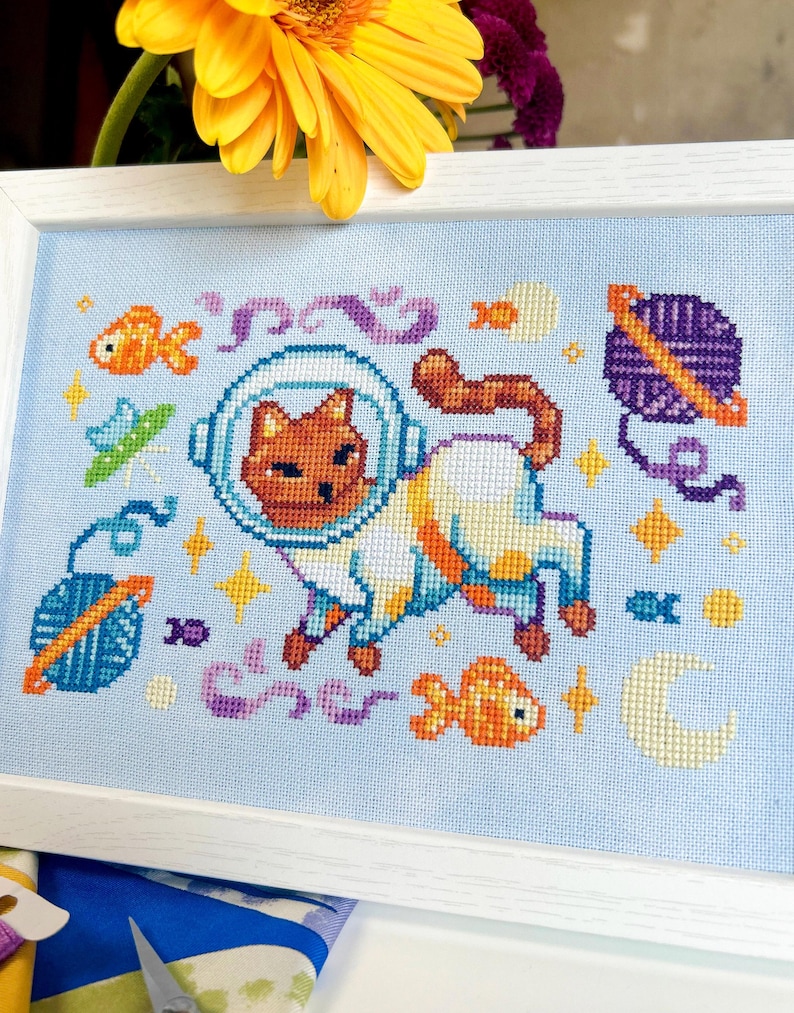 May include: Cross-stitch art featuring a cat in an astronaut suit, surrounded by space-themed elements like planets, fish, and stars. The artwork is set against a light blue background and framed in a white wooden frame. The cat is brown and white.