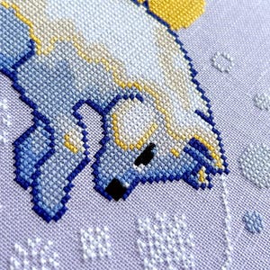 May include: Close-up of a cross-stitch pattern of a white polar bear with blue and yellow accents, on a purple background with white snowflakes.