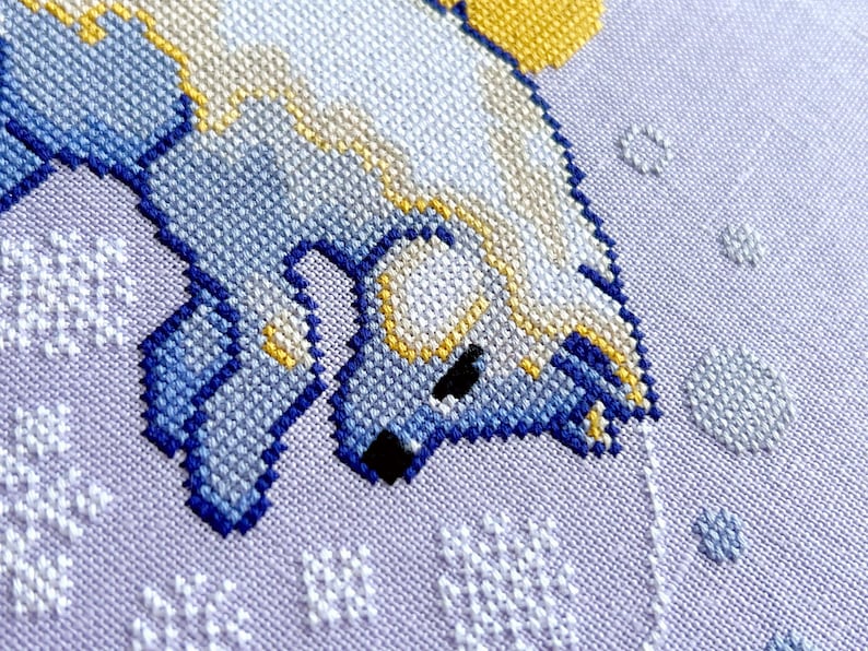 May include: Close-up of a cross-stitch pattern of a white polar bear with blue and yellow accents, on a purple background with white snowflakes.