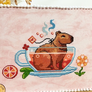 K&ouml;nnte beinhalten: Ein Kreuzstichmuster eines Kapibaras, das in einer Teetasse mit einer dampfenden Tasse Tee sitzt. Das Kapibara ist braun und die Teetasse ist blau mit einem wei&szlig;en Unterteller. Im Hintergrund befinden sich eine kleine Orangenscheibe und eine Blume.