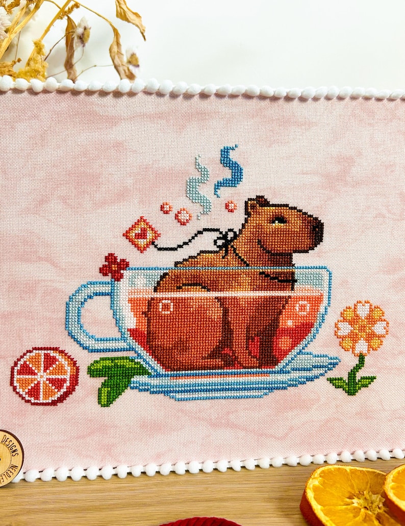 K&ouml;nnte beinhalten: Ein Kreuzstichmuster eines Kapibaras, das in einer Teetasse mit einer dampfenden Tasse Tee sitzt. Das Kapibara ist braun und die Teetasse ist blau mit einem wei&szlig;en Unterteller. Im Hintergrund befinden sich eine kleine Orangenscheibe und eine Blume.