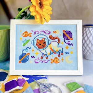 May include: Cross-stitch art of a cat in a space helmet, surrounded by fish, stars, and planets, framed in a white wooden frame. The artwork is on a light blue background. A yellow flower and spools of thread are in the background.