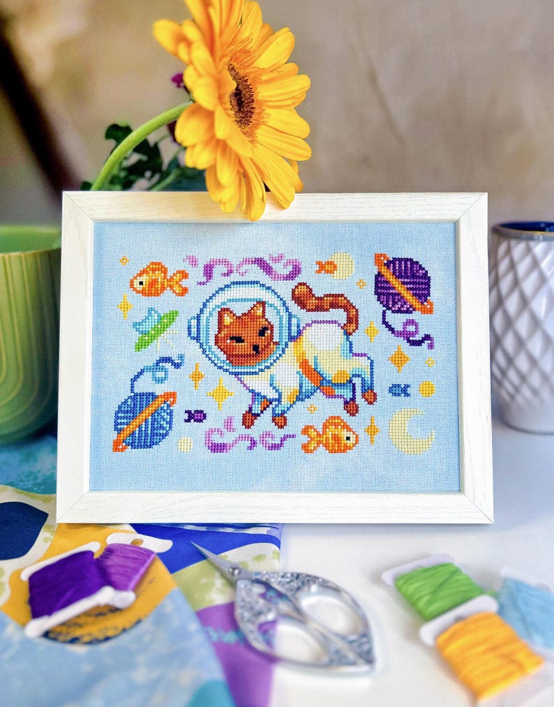 May include: Cross-stitch art of a cat in a space helmet, surrounded by fish, stars, and planets, framed in a white wooden frame. The artwork is on a light blue background. A yellow flower and spools of thread are in the background.