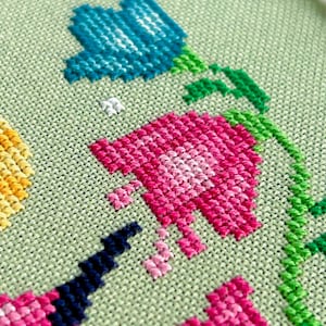 May include: Close-up of a cross-stitch floral design featuring blue and pink flowers with green stems and leaves. The flowers are stitched on a light green fabric, showcasing the detailed needlework.