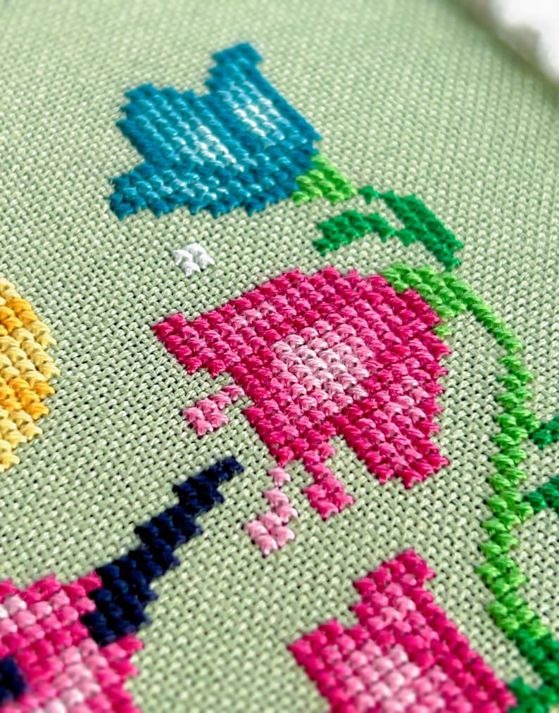 May include: Close-up of a cross-stitch floral design featuring blue and pink flowers with green stems and leaves. The flowers are stitched on a light green fabric, showcasing the detailed needlework.