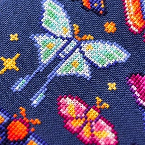 Op de afbeelding: Close-up van een kruissteekontwerp met kleurrijke vlinders en stervormige accenten op een donkerblauwe stof. De vlinders zijn uitgevoerd in verschillende kleuren, waaronder blauw, oranje, roze en groen, met gedetailleerde vleugelpatronen.