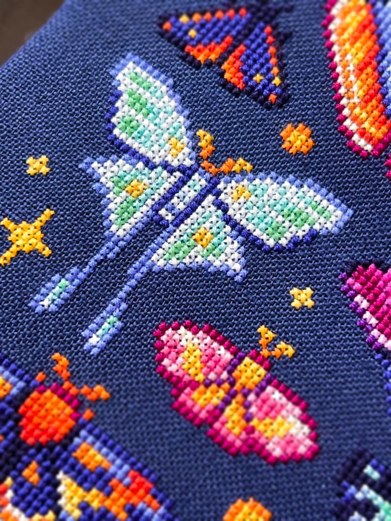 Op de afbeelding: Close-up van een kruissteekontwerp met kleurrijke vlinders en stervormige accenten op een donkerblauwe stof. De vlinders zijn uitgevoerd in verschillende kleuren, waaronder blauw, oranje, roze en groen, met gedetailleerde vleugelpatronen.