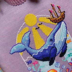 Closeup of Whale with Boat cross stitch pattern. Stitches are neat and tidy. There are little detailsin the wale, like shimmers. The boat is brown with sails and pink flags. The sun rays permeate the water. Bubbles are placed on the sides.