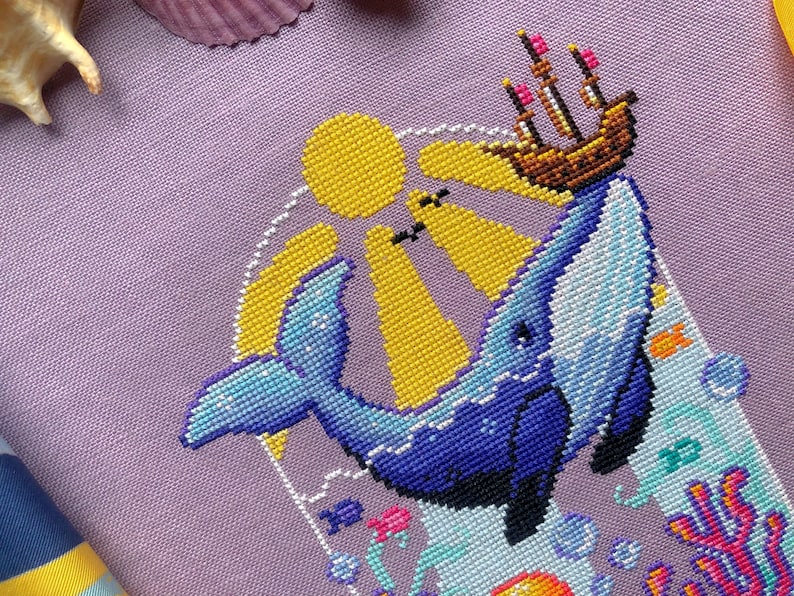 Closeup of Whale with Boat cross stitch pattern. Stitches are neat and tidy. There are little detailsin the wale, like shimmers. The boat is brown with sails and pink flags. The sun rays permeate the water. Bubbles are placed on the sides.