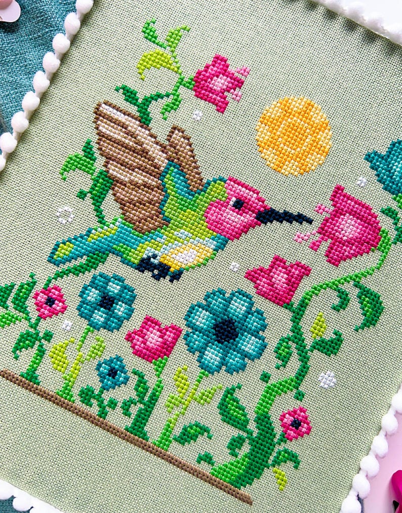 May include: Cross-stitch embroidery of a hummingbird in flight, surrounded by flowers and foliage. The bird has green, blue, and pink coloring, with brown wings. The flowers are pink and blue, with a yellow sun in the background. The piece is framed with white pom-poms.