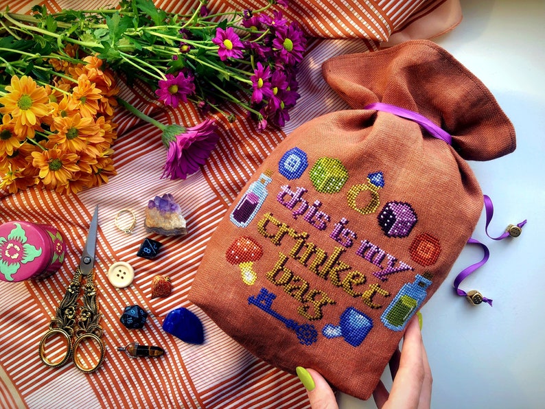Trinket bag cross stitch front view. The phrase This is my Trinket Bag is stitched onto a drawstring bag. It is surrounded by objects, such as dice, potion bottles, mushrooms, buttons, a key and a ring. The bag is brown. The scene is very colorful.
