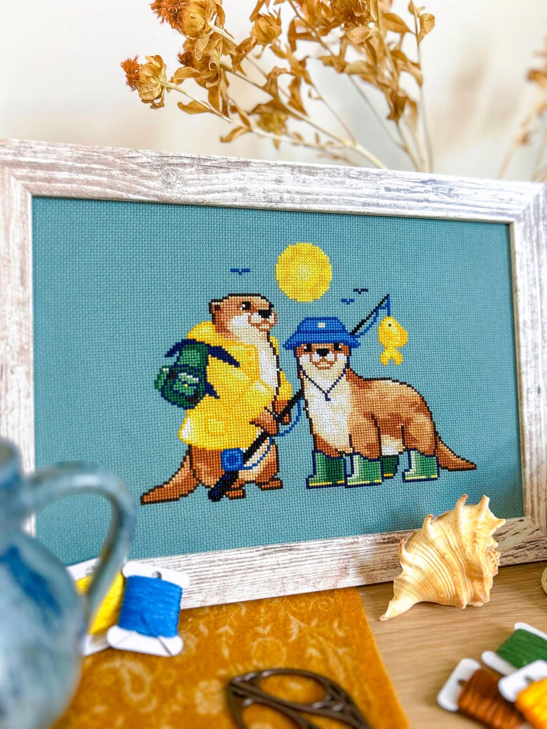 May include: Cross-stitch artwork in a wooden frame. The image shows two otters: one in a yellow raincoat and backpack, the other fishing with a blue hat and wellington boots. A yellow sun and blue background complete the scene.