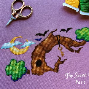 May include: Cross stitch pattern of a brown tree with a key hanging from a branch, a crescent moon, and three green shamrocks. The text "The Secret Grove Part 1" is stitched below the tree.