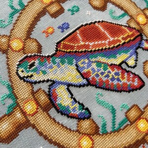 May include: A colorful cross-stitch design of a sea turtle swimming in the ocean. The turtle is surrounded by a wooden ship's wheel and seaweed. The turtle has a red, orange, yellow, green, blue, and purple shell.