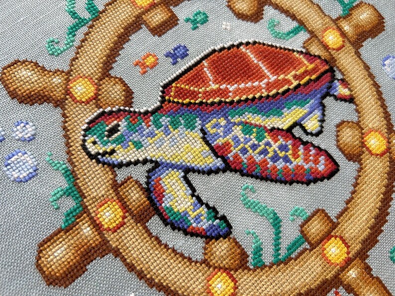 May include: A colorful cross-stitch design of a sea turtle swimming in the ocean. The turtle is surrounded by a wooden ship's wheel and seaweed. The turtle has a red, orange, yellow, green, blue, and purple shell.