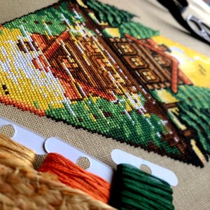 Closeup of the Lakeside Cabin cross stitch pattern. Image is zoomed in on the lower end of the design, where the water reflection is visible. The colors are vibrant, the stitches are neat and uniform. In the forefront are the rim of a hat and bobbins