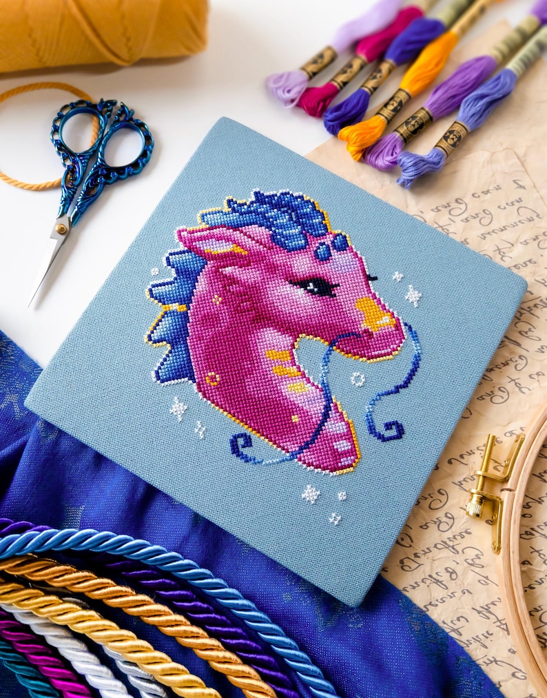 Pode incluir: Um projeto de ponto cruz conclu&iacute;do com uma cabe&ccedil;a de drag&atilde;o rosa e azul em tecido azul claro. A imagem inclui fios de bordar em v&aacute;rias cores, tesouras azuis e um bastidor de bordado, sugerindo um tema de artesanato.
