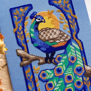 May include: A cross-stitch embroidery on light blue fabric featuring a peacock perched on a brown branch. The peacock has blue, green, and brown feathers with a long tail. A yellow sun is behind the bird, framed by blue and gold decorative borders.