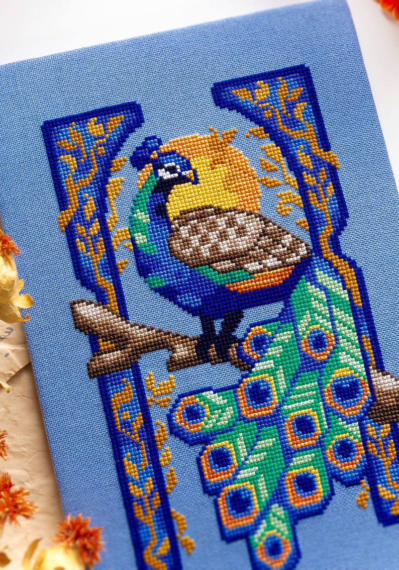May include: A cross-stitch embroidery on light blue fabric featuring a peacock perched on a brown branch. The peacock has blue, green, and brown feathers with a long tail. A yellow sun is behind the bird, framed by blue and gold decorative borders.