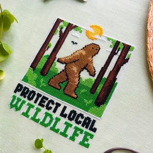 May include: Cross-stitch art featuring a brown Bigfoot walking through a forest scene with a yellow sun. The text "PROTECT LOCAL WILDLIFE" is stitched in green and black. The piece is surrounded by green fabric and embroidery floss.