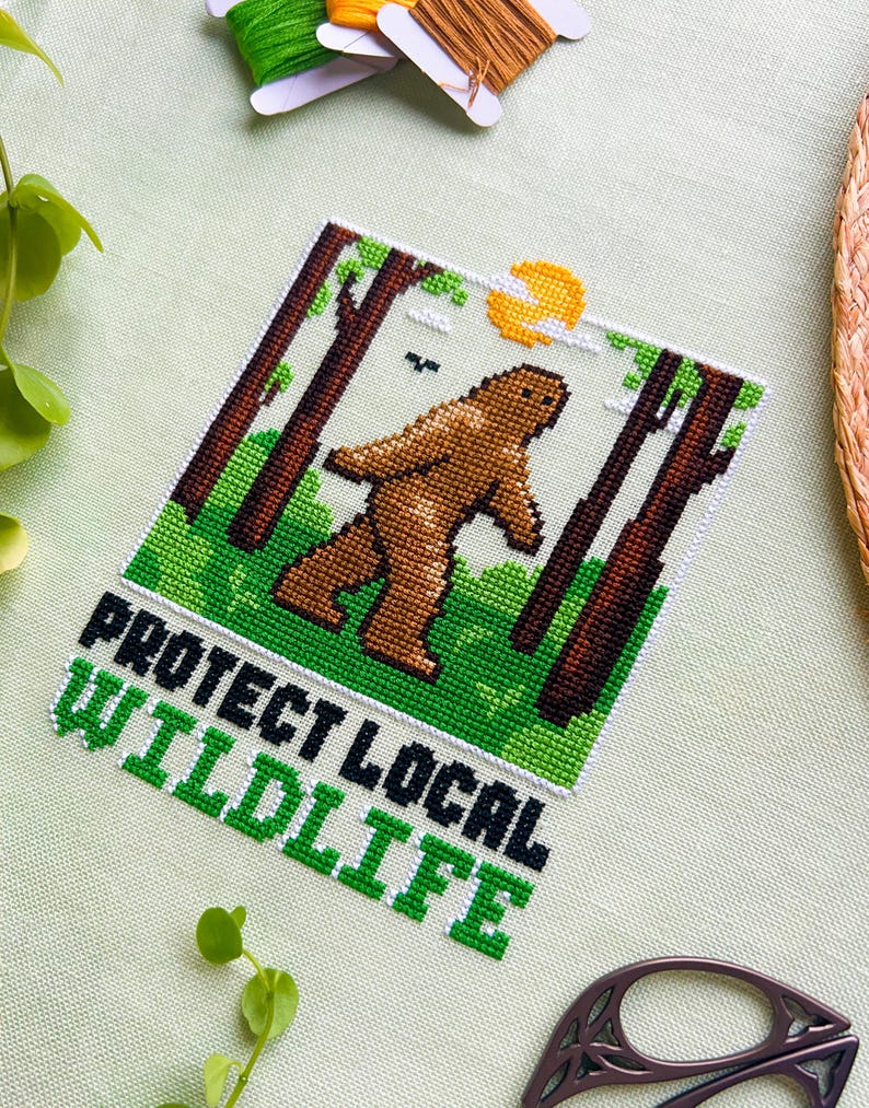 May include: Cross-stitch art featuring a brown Bigfoot walking through a forest scene with a yellow sun. The text "PROTECT LOCAL WILDLIFE" is stitched in green and black. The piece is surrounded by green fabric and embroidery floss.