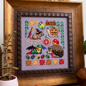 May include: Cross-stitch artwork in a wooden frame. The design features Halloween-themed elements: a rat with a witch hat, a pumpkin, a snail, a frog, and a hedgehog. The background is lavender, with autumn leaves and the text "OCT31".