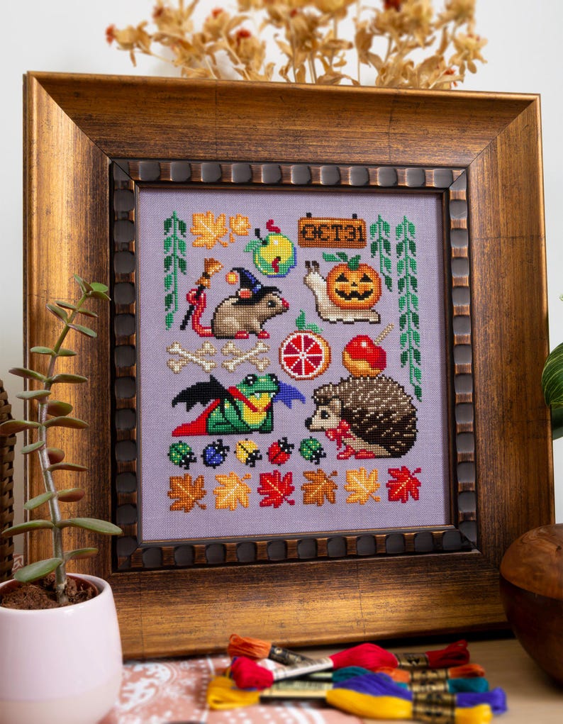 May include: Cross-stitch artwork in a wooden frame. The design features Halloween-themed elements: a rat with a witch hat, a pumpkin, a snail, a frog, and a hedgehog. The background is lavender, with autumn leaves and the text "OCT31".