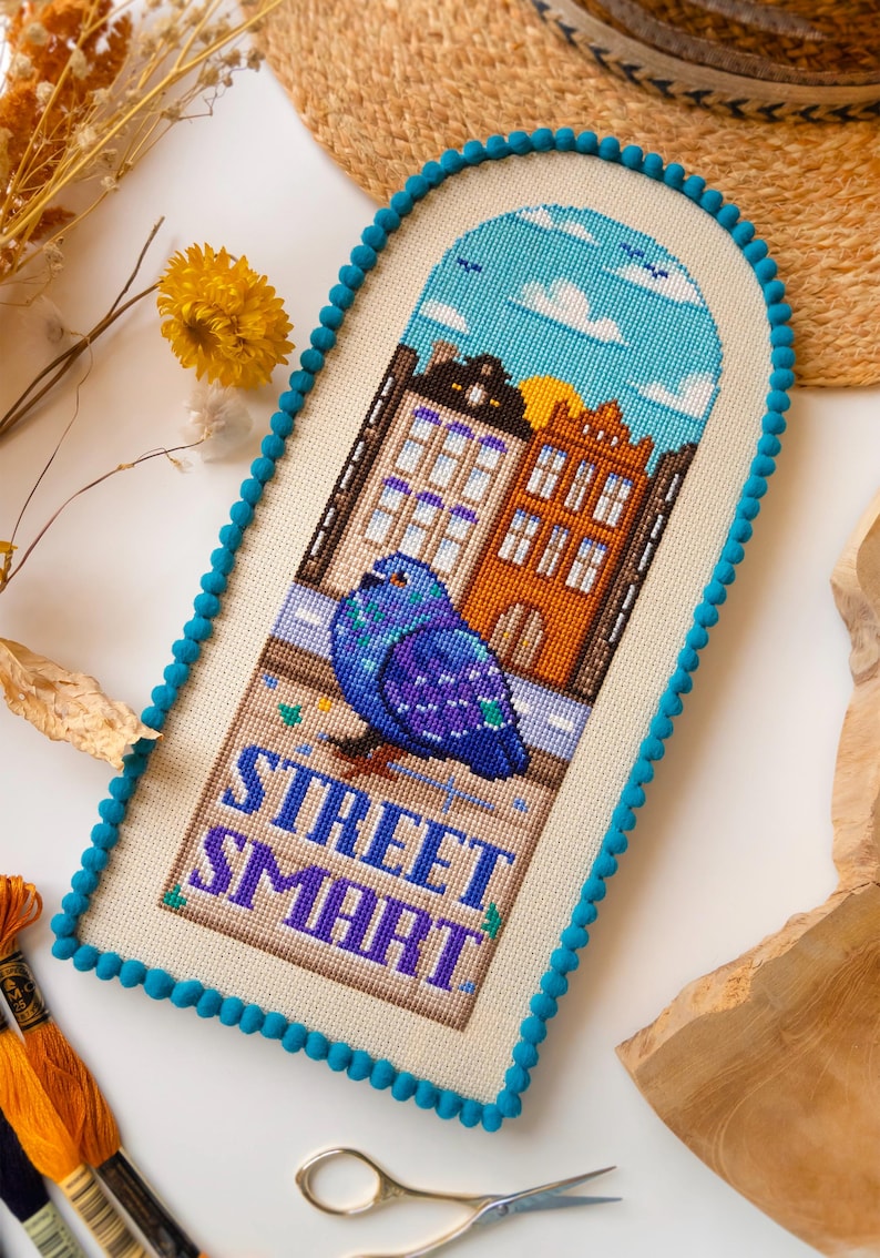 Puede incluir: Una obra de arte de punto de cruz que representa una paloma azul frente a edificios, con el texto "STREET SMART". La obra de arte tiene un borde de pomp&oacute;n azul y un cielo azul con nubes. Los edificios son naranjas y morados. La obra est&aacute; enmarcada.