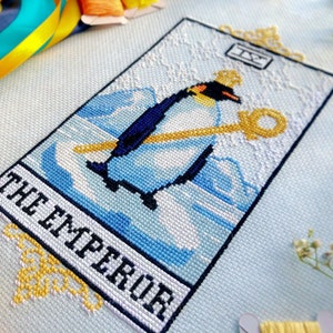 Closeup of the Emperor tarot cross stitch pattern. Image is focused on the lower right corner of the design. Emperor penguin is standing proudly in his attire on a block of ice, surrounded by water and icebergs. The frame is that of a tarot card.