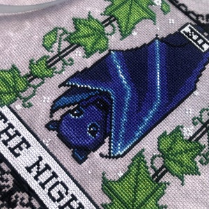 May include: Cross-stitch embroidery of a bat with blue wings, hanging from a vine with green leaves. The text "OF THE NIGHT" is stitched below the bat.