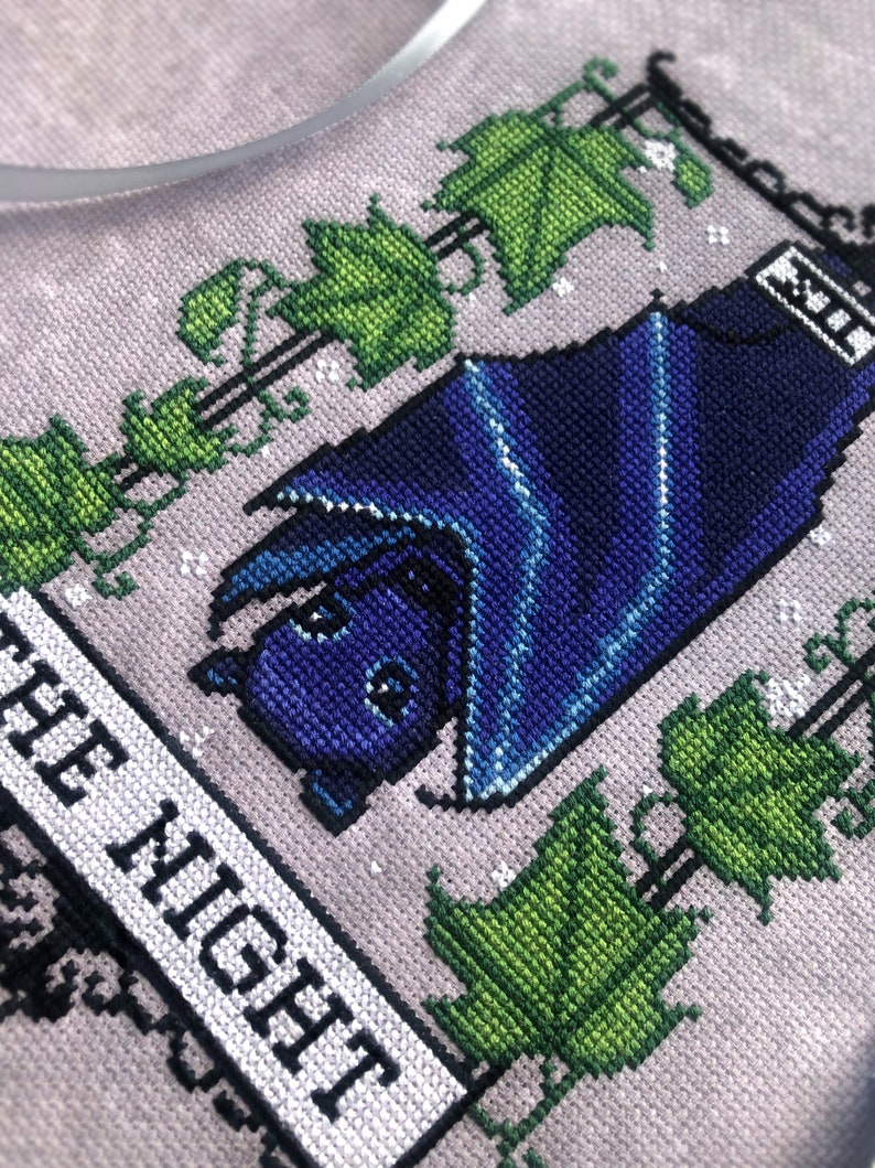 May include: Cross-stitch embroidery of a bat with blue wings, hanging from a vine with green leaves. The text "OF THE NIGHT" is stitched below the bat.