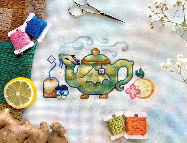 May include: A cross-stitch pattern of a green teapot with a dragon handle, surrounded by lemons, flowers, and tea bags. The pattern is on a light blue background with white clouds.