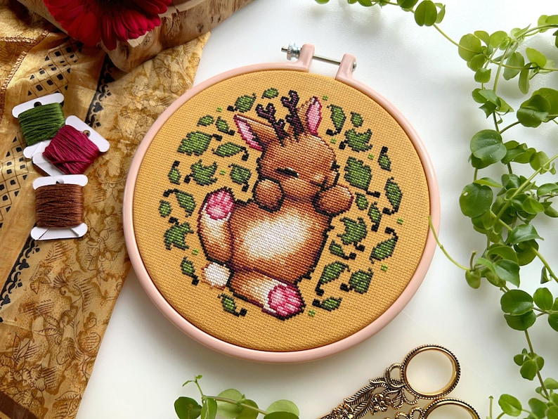 May include: A cross-stitch embroidery hoop with a light brown deer with antlers curled up in a circle of green leaves on a yellow background.