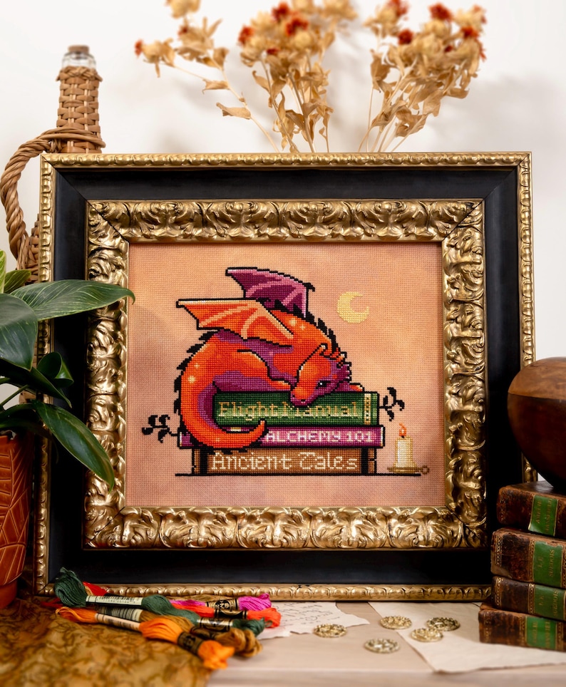 May include: Cross-stitch art of a red and purple dragon resting on books titled "Flight Manual", "Alchemy 101", and "Ancient Tales". The framed artwork has a gold ornate frame and a black inner frame. A small candle is lit.
