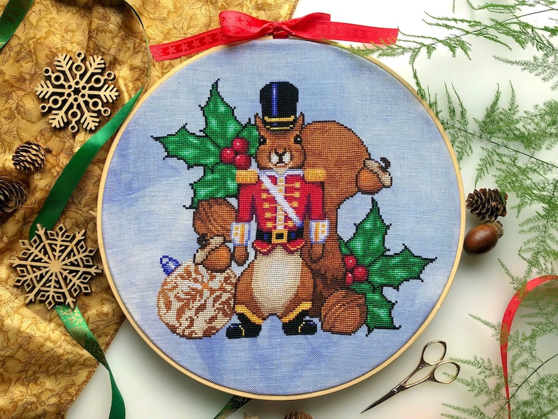 Front view of Nutcracker squirrel cross stitch pattern. Squirrel is wearing a military suit and looks stern. His jacket is red and he is wearing black boots and a hat. Around him are walnuts, acorns, holly and a bauble. Scene is surrounded by decor.