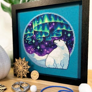 May include: A framed cross-stitch design featuring a polar bear gazing up at a vibrant green, purple, and blue aurora borealis against a dark blue background.