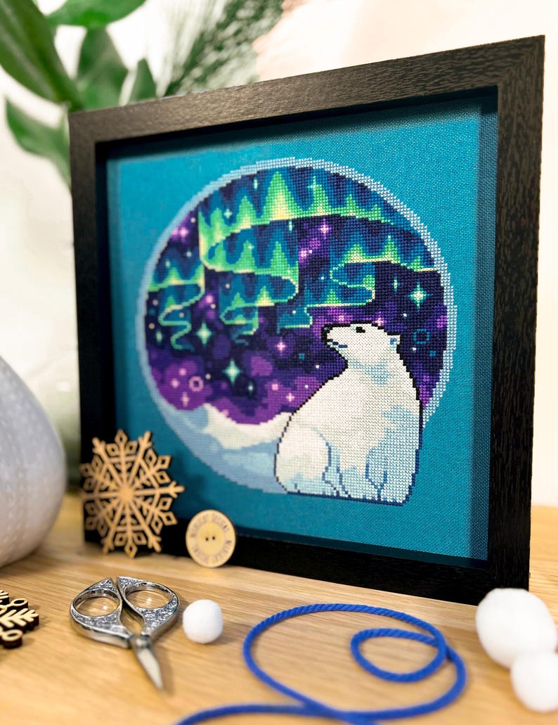 May include: A framed cross-stitch design featuring a polar bear gazing up at a vibrant green, purple, and blue aurora borealis against a dark blue background.