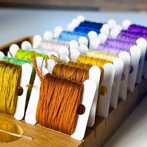 May include: A wooden organizer filled with spools of colorful embroidery floss. The floss is arranged in a rainbow of colors, including yellow, orange, green, blue, and purple. Each spool is neatly wound and held in place by a white card.